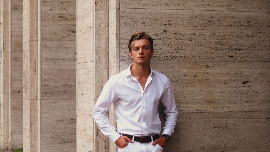 A young man in a white shirt leans against a modern architectural wall.